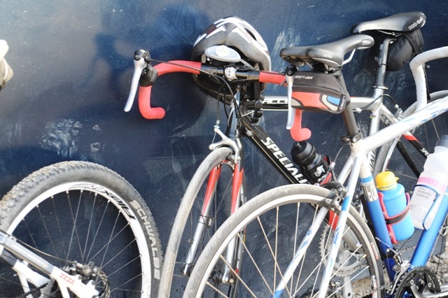 Fotografia de bicicletas encostadas a uma parede com os capacetes e garrafas de água